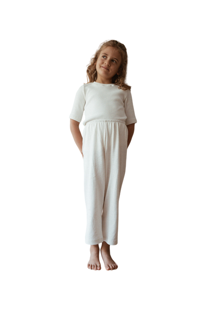 A young girl with wavy blonde hair stands barefoot, wearing a white outfit that includes a short-sleeved top and loose-fitting Resort Pant in Hemp by cabane childrenswear. Ethically handmade in Calgary, she is looking upwards to her left with a thoughtful, slight smile, her hands loosely clasped behind her back.