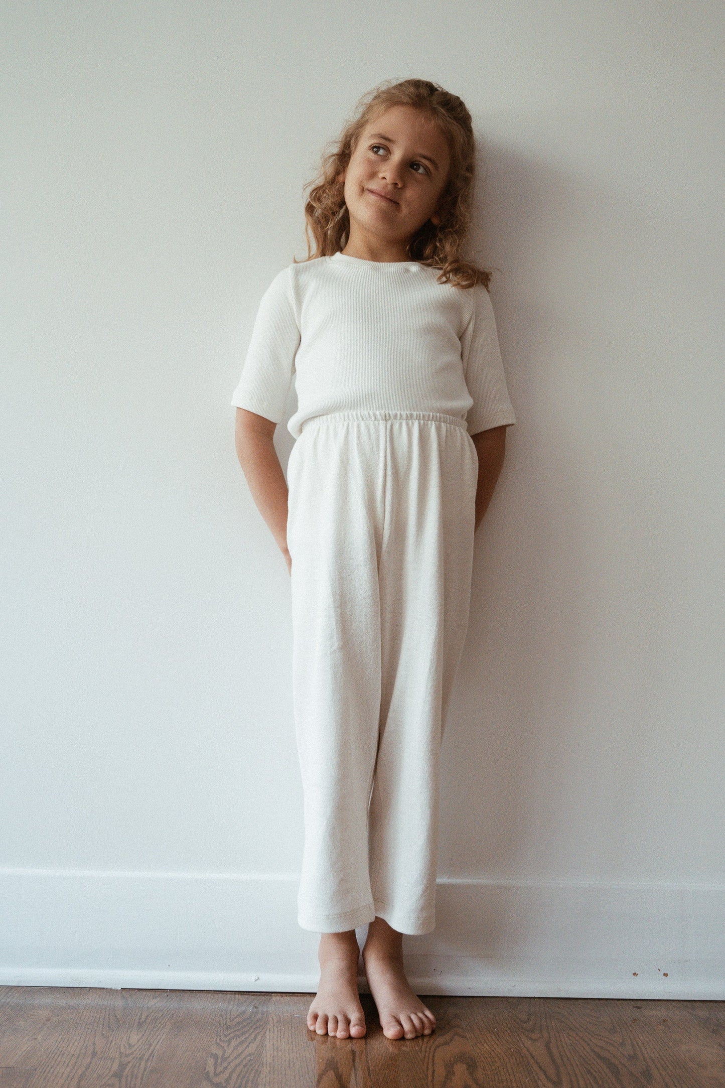 A young child with wavy, shoulder-length hair stands barefoot against a plain white wall. Dressed in a white, short-sleeved top and ethically handmade RESORT PANT hemp from cabane childrenswear, they look slightly to the side with a small, thoughtful smile. The floor is wooden.