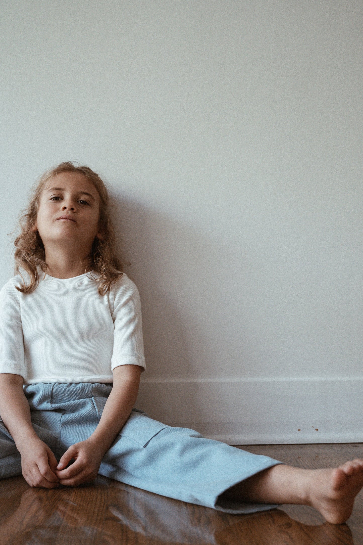 A child with curly hair sits on the floor against a plain wall, dressed in a Classic Top for Baby + Kid by cabane childrenswear, paired with light blue pants. One leg is extended and barefoot, and the child's expression is relaxed.