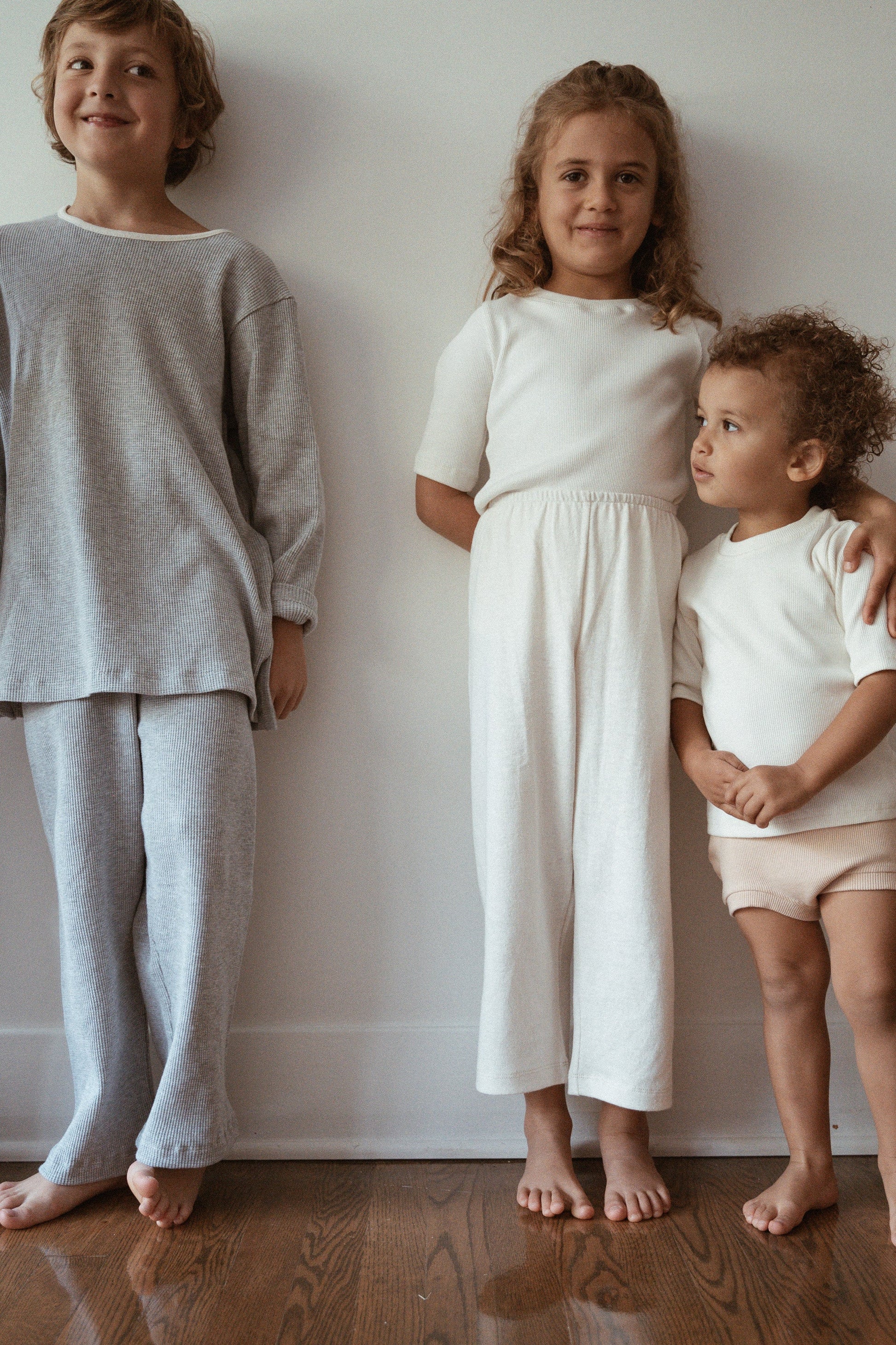 Three children stand barefoot indoors against a white wall. The child on the left wears a gray 2x2 rib outfit, the child in the middle wears a white dress, and the child on the right sports an ethically handmade CLASSIC TOP baby + kid by cabane childrenswear with beige shorts. They all have light brown curly hair.