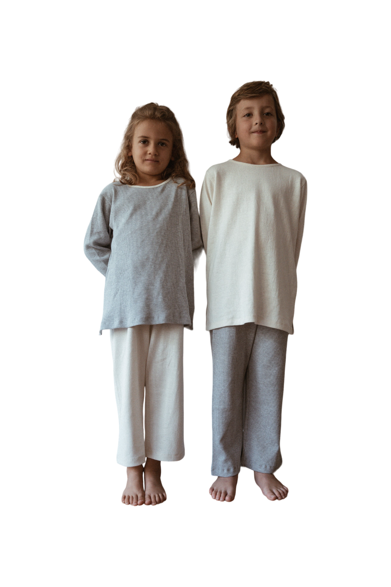 Two children are standing side by side against a plain background, both barefoot. The child on the left is wearing a grey organic cotton long-sleeve top and white pants, while the child on the right sports a white handmade in Calgary RESORT TOP hemp jersey by cabane childrenswear along with grey pants. Both are smiling slightly.