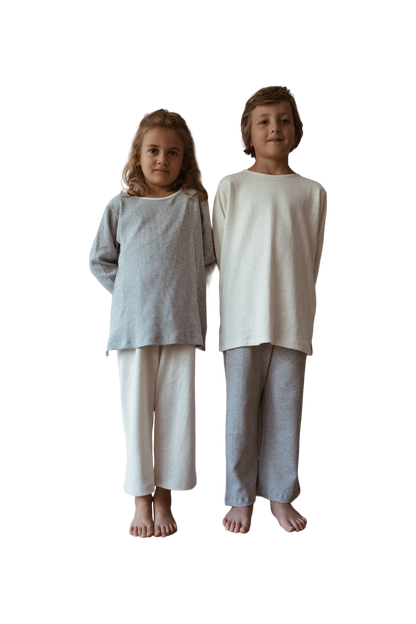 Two children are standing side by side against a plain background, both barefoot. The child on the left is wearing a grey organic cotton long-sleeve top and white pants, while the child on the right sports a white handmade in Calgary RESORT TOP hemp jersey by cabane childrenswear along with grey pants. Both are smiling slightly.