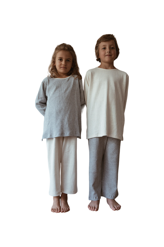 Two children are standing side by side against a plain background, both barefoot. The child on the left is wearing a grey organic cotton long-sleeve top and white pants, while the child on the right sports a white handmade in Calgary RESORT TOP hemp jersey by cabane childrenswear along with grey pants. Both are smiling slightly.