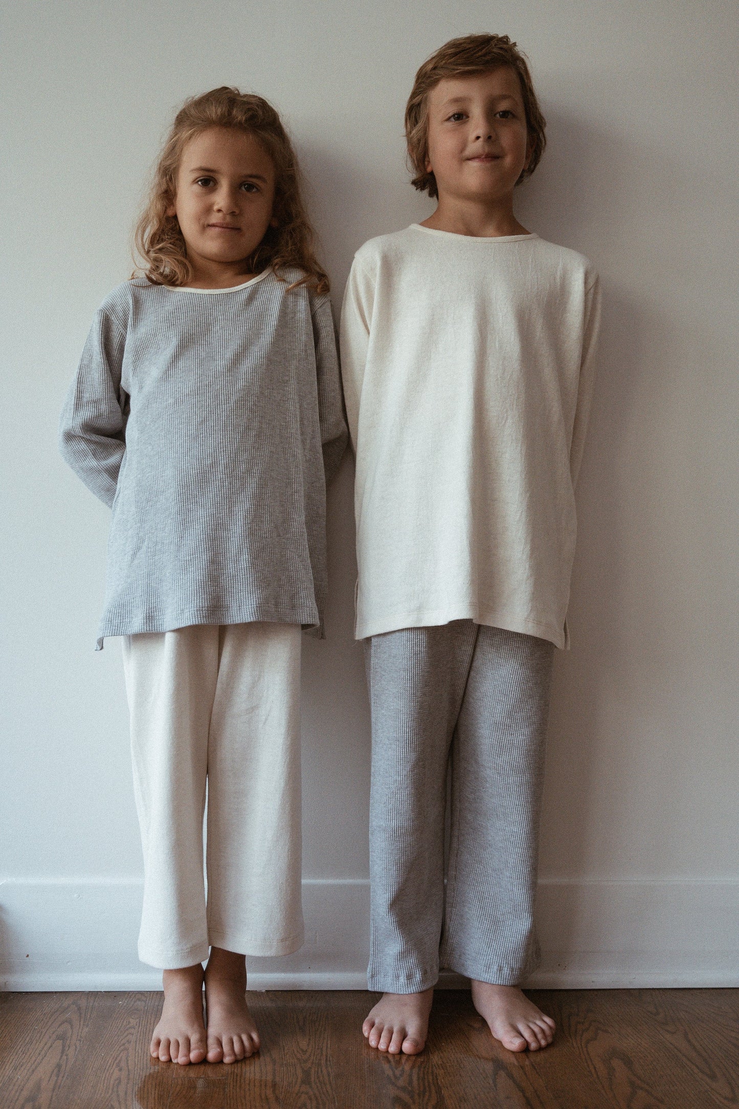 Two children stand side by side against a plain wall, both dressed in comfortable, loose-fitting, neutral-toned pajamas handmade in Calgary. The child on the left is wearing an organic cotton light gray top and cream-colored pants, while the child on the right sports a cream-colored RESORT TOP hemp jersey from cabane childrenswear paired with light gray pants. Both children are barefoot.