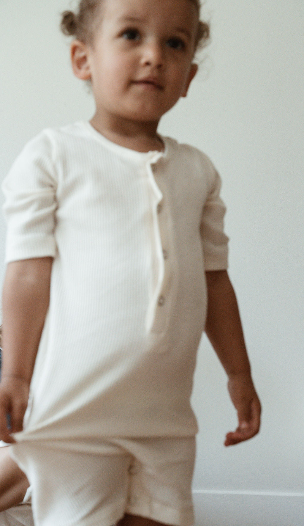 A young child with light brown hair stands wearing the cabane childrenswear pre-order HIGH-NOON SUNSUIT, a white, ribbed onesie made from undyed organic cotton and featuring front buttons. The background is simply decorated and softly lit.