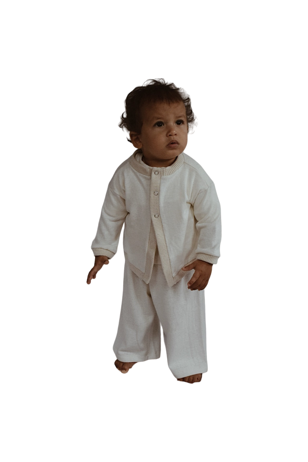 A toddler with curly hair stands barefoot, dressed in a white, ethically handmade Soul Sweater Baby + Kid and matching loose-fitting pants made from organic cotton and hemp by cabane childrenswear, looking slightly to the side.