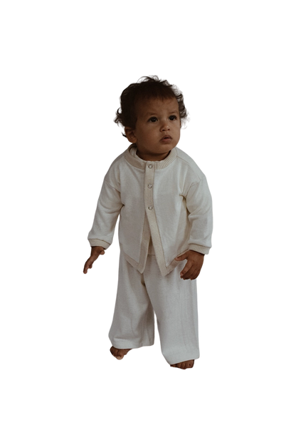 A toddler with curly hair stands barefoot, dressed in a white, ethically handmade Soul Sweater Baby + Kid and matching loose-fitting pants made from organic cotton and hemp by cabane childrenswear, looking slightly to the side.