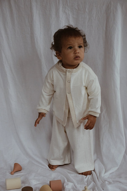 A young child stands against a white fabric backdrop, looking upward with a thoughtful expression. The child is wearing the cozy, cream-colored Soul Sweater Baby + Kid from cabane childrenswear, made of organic cotton and hemp and featuring a boxy design with 3 snaps. Matching pants complete the ensemble, while various wooden toys are scattered nearby.