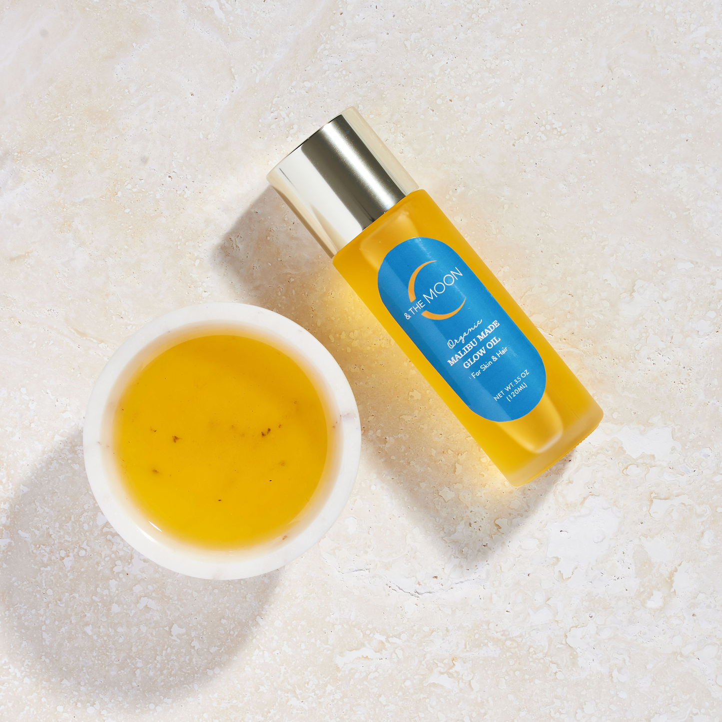 A small white bowl filled with a yellow-orange liquid sits on a textured beige surface. Next to the bowl is a cylindrical bottle adorned with a blue label and gold cap, featuring the text "Malibu Made Glow Oil" at the top and "C & The Moon" in the middle. This serene scene embodies an ideal clean skincare routine.