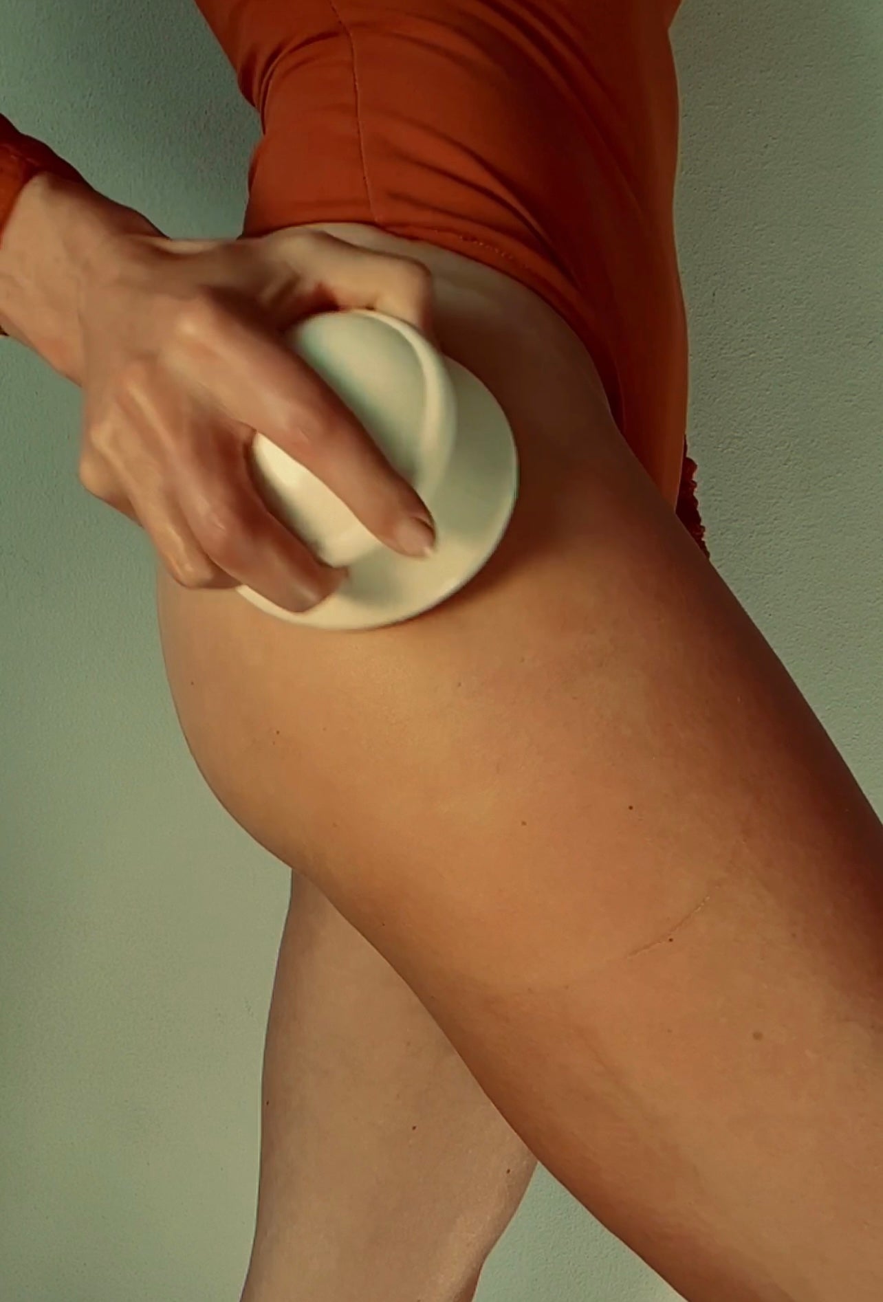 A person dressed in an orange garment is using LENI's The Face & Body Cupping Set, specifically the white cupping massage tool, on their thigh, concentrating on the upper leg. The background is blurred to accentuate the action of applying this tool, underscoring its benefits for promoting a healthy lymphatic system.