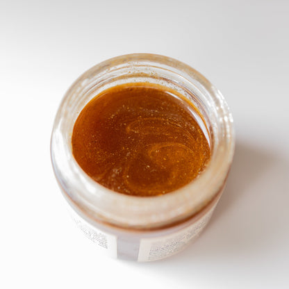 An open jar of Honeybee Hippie's Herbal Infused Raw Honey Mask, a thick, amber-colored substance, glistens against the white background. Its smooth surface highlights its purity and potential detoxifying benefits.