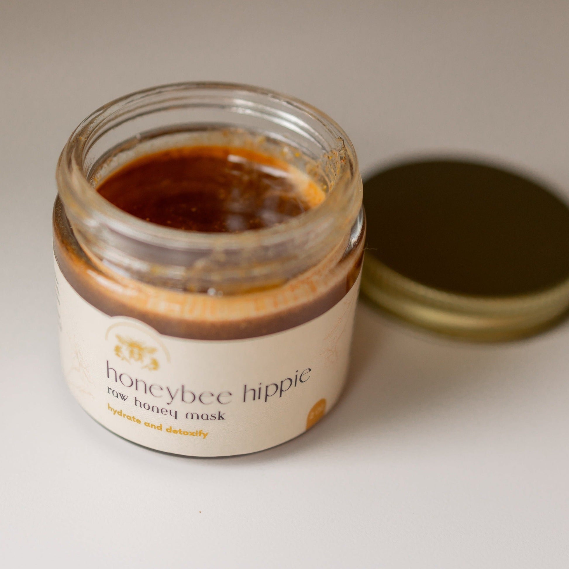 An open glass jar of Honeybee Hippie's herbal infused raw honey mask, with the lid set aside, reveals a thick, brown mask promising hydration and detoxification. The label reads "hydration and detoxify," assuring skin rejuvenation.