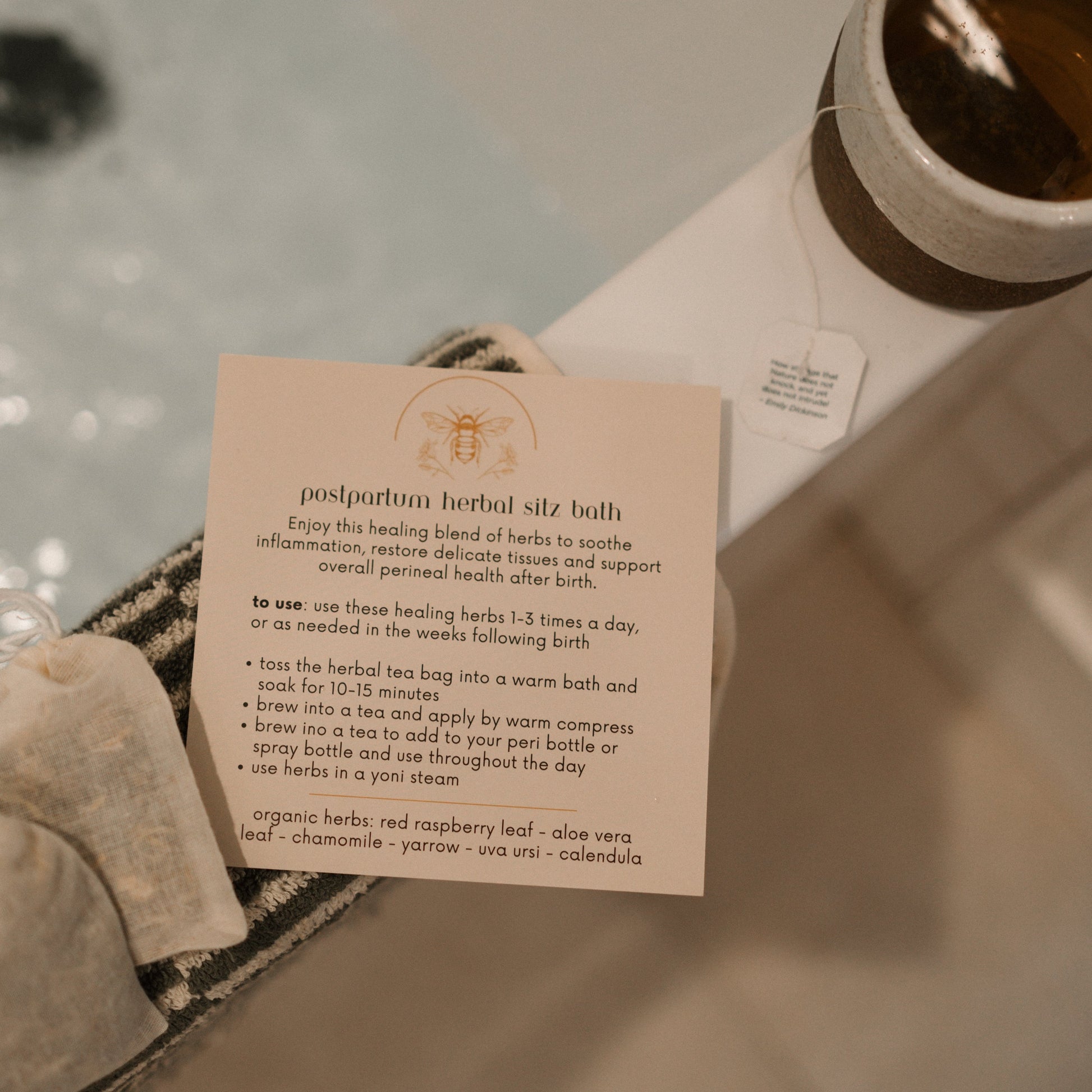 A Honeybee Hippie postpartum herbal sitz bath instruction card sits on a towel beside the bathtub, describing how to use herbal tea bags with raspberry leaf, aloe vera, and chamomile for perineal healing. A teacup rests on the tub's edge for soothing relief for postpartum moms.