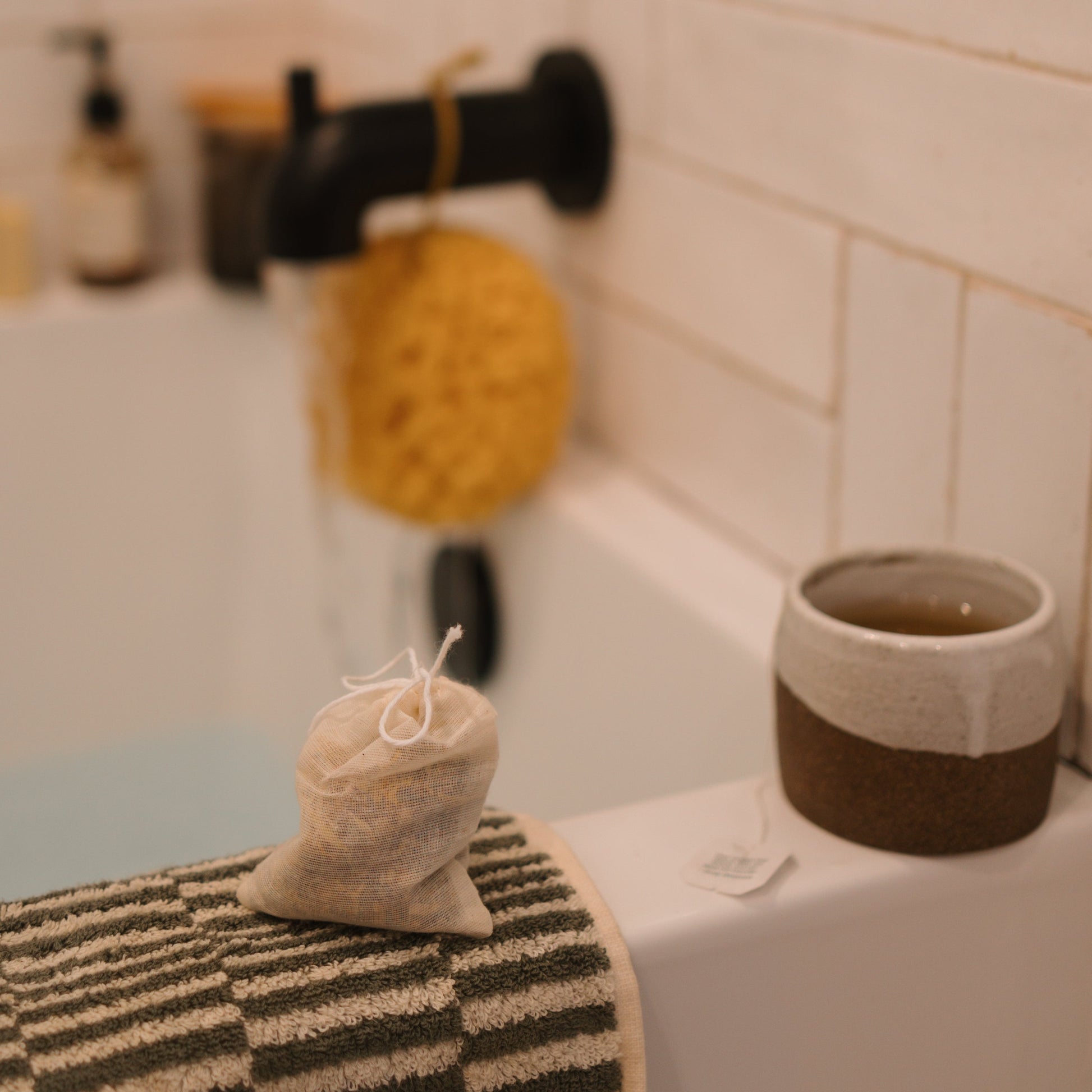 A cozy bathroom features a close-up of a bathtub corner with a striped towel draped over the edge, topped with Honeybee Hippie's postpartum herbal sitz bath bag. Nearby, a mug with the tea bag for soothing and a yellow sponge hang from the wall.