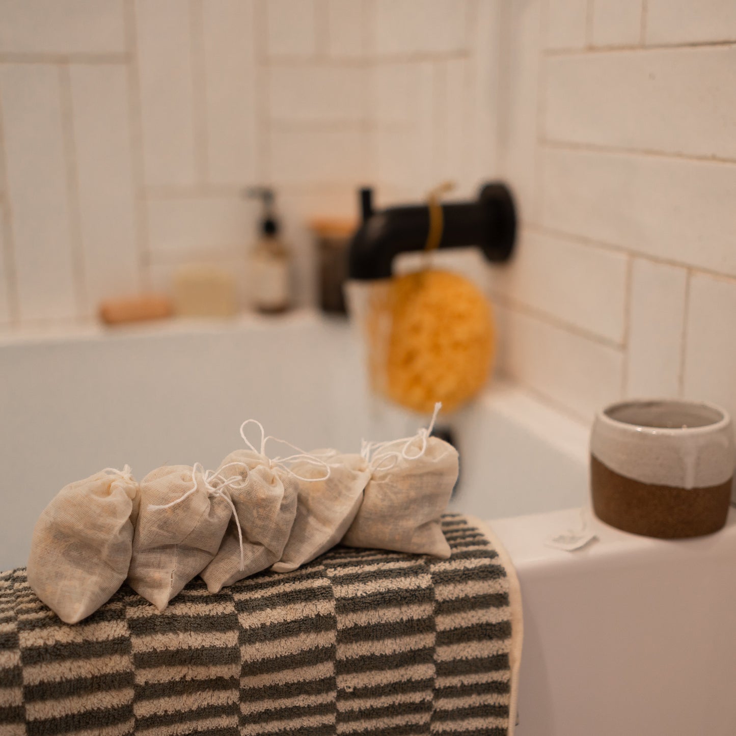 In a bathroom setting, four Honeybee Hippie postpartum herbal sitz bath bags are lined up on a striped towel draped over the bathtub edge. A yellow sponge and pump bottle rest near the black faucet, while a ceramic cup sits on the tub's rim.