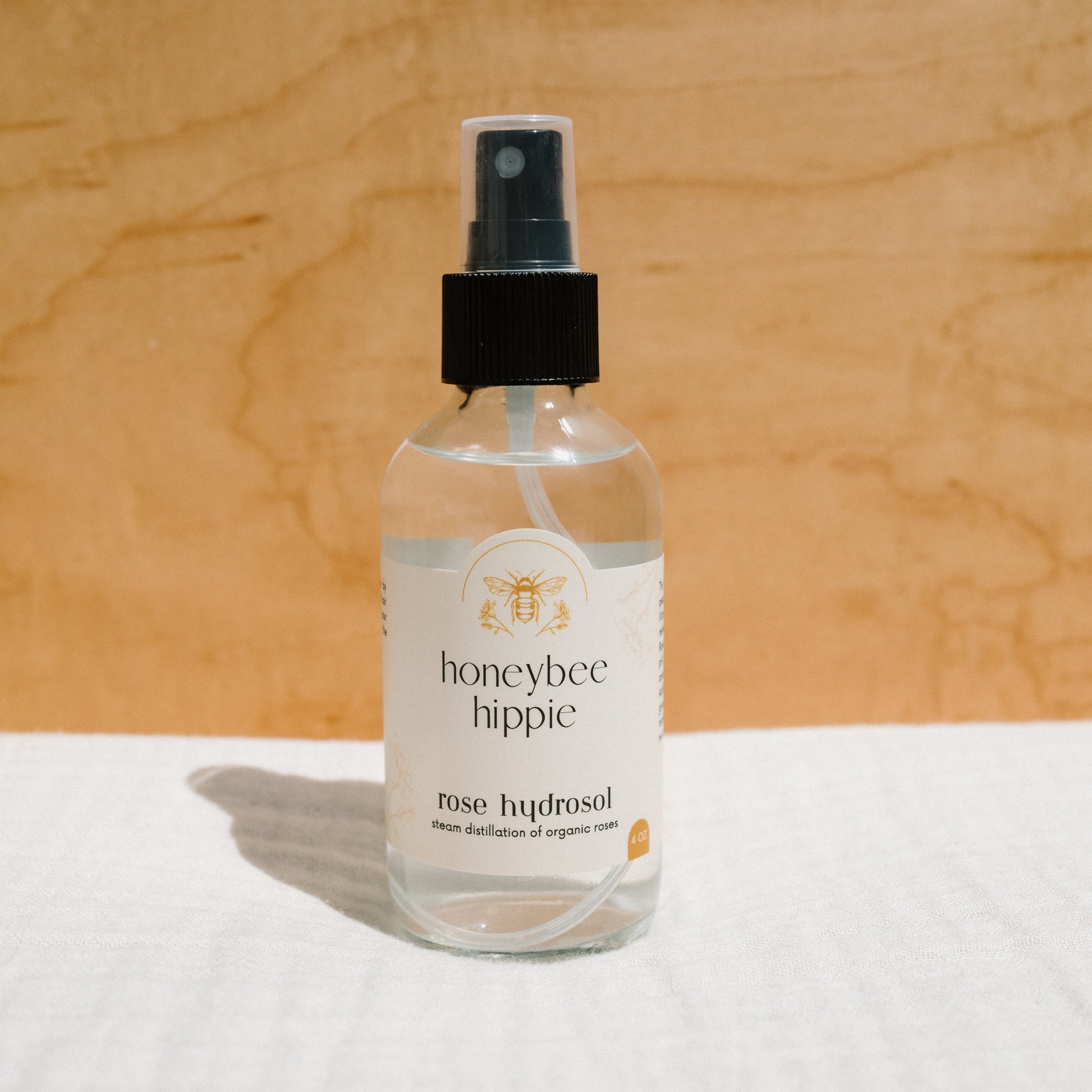 A Honeybee Hippie rose hydrosol spray bottle rests on a light surface with a wooden backdrop. Its label features a bee and describes it as "steam distillation of organic roses," ideal for enhancing your skincare regimen.