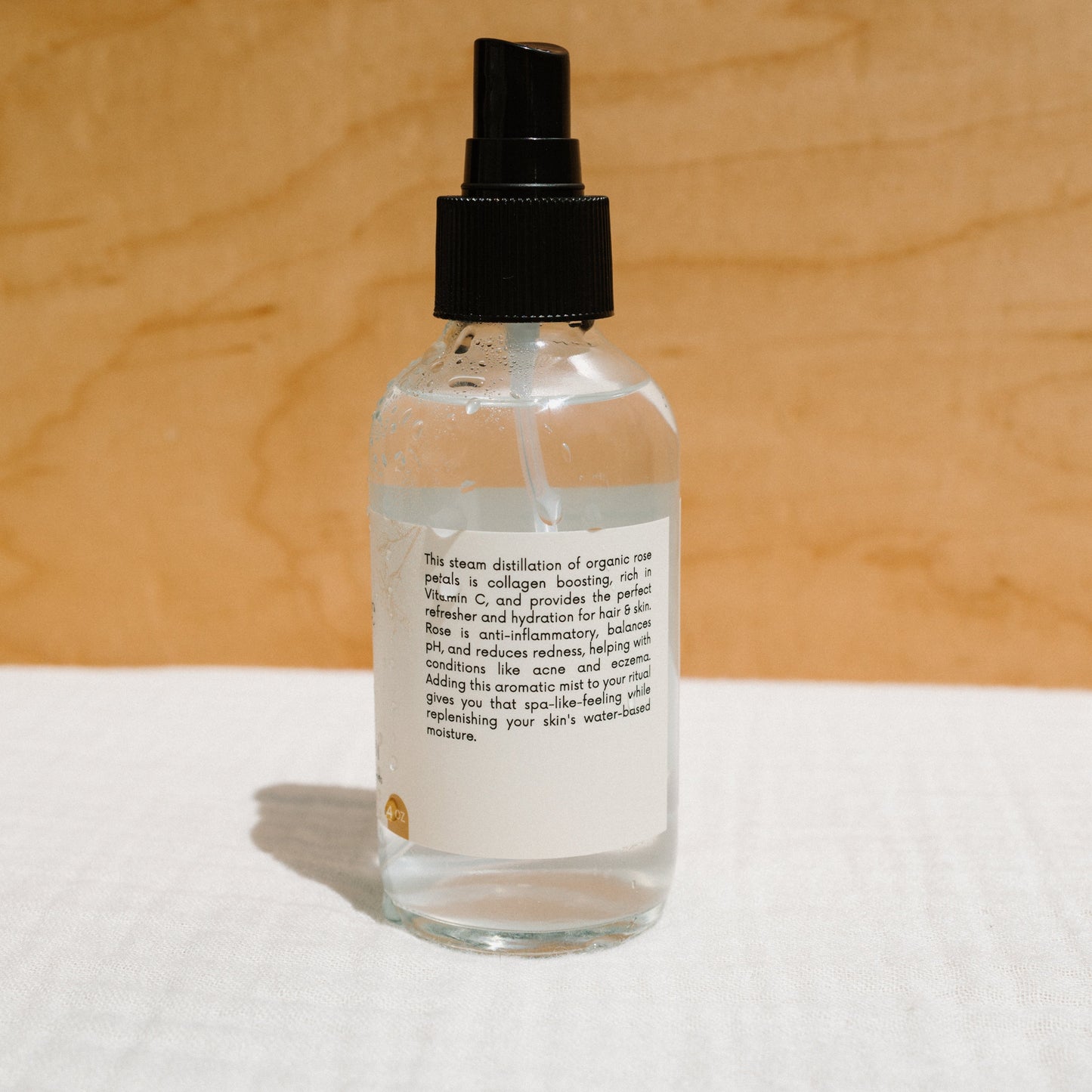 A glass spray bottle with a clear liquid sits on a white surface against a wooden background. The blurred label suggests the benefits of Honeybee Hippie's Rose Hydrosol, promising to enhance your skincare routine. It features a black spray nozzle, ready to refresh and moisturize your skin.