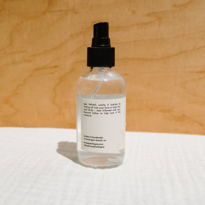 A clear spray bottle with a black nozzle labeled "Honeybee Hippie Rose Hydrosol" rests on light brown wood, indicating it's an organic rose petal face mist with usage instructions and contact info.