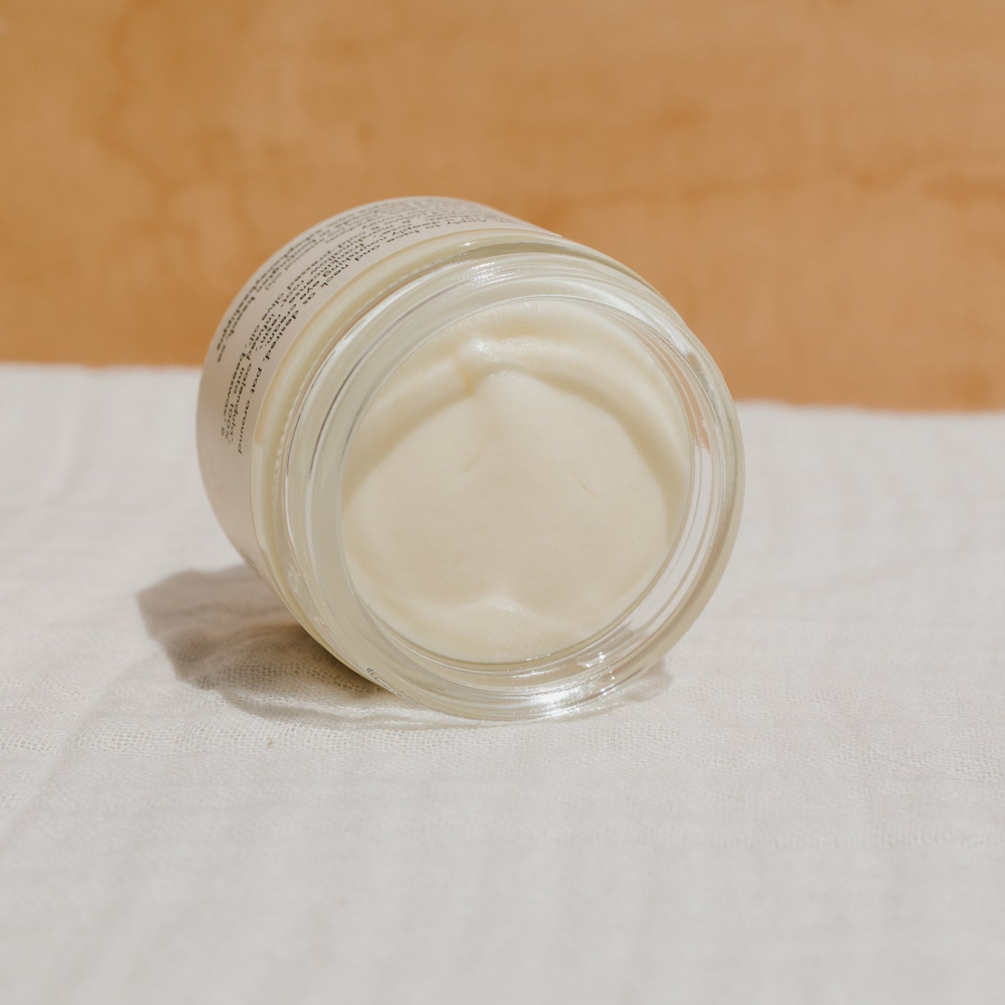 A glass jar labeled "honey hydrate whipped tallow butter" by Honeybee Hippie, tipped on its side with creamy contents inside, rests on a textured white surface. A warm, light brown backdrop enhances the inviting scene.