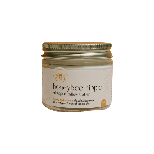 The scene showcases a jar of Honeybee Hippie's "honey hydrate whipped tallow butter," featuring a golden lid against a sleek black background. Enhanced with Manuka honey, the product is suitable for all skin types and promises nourishment for aging skin.