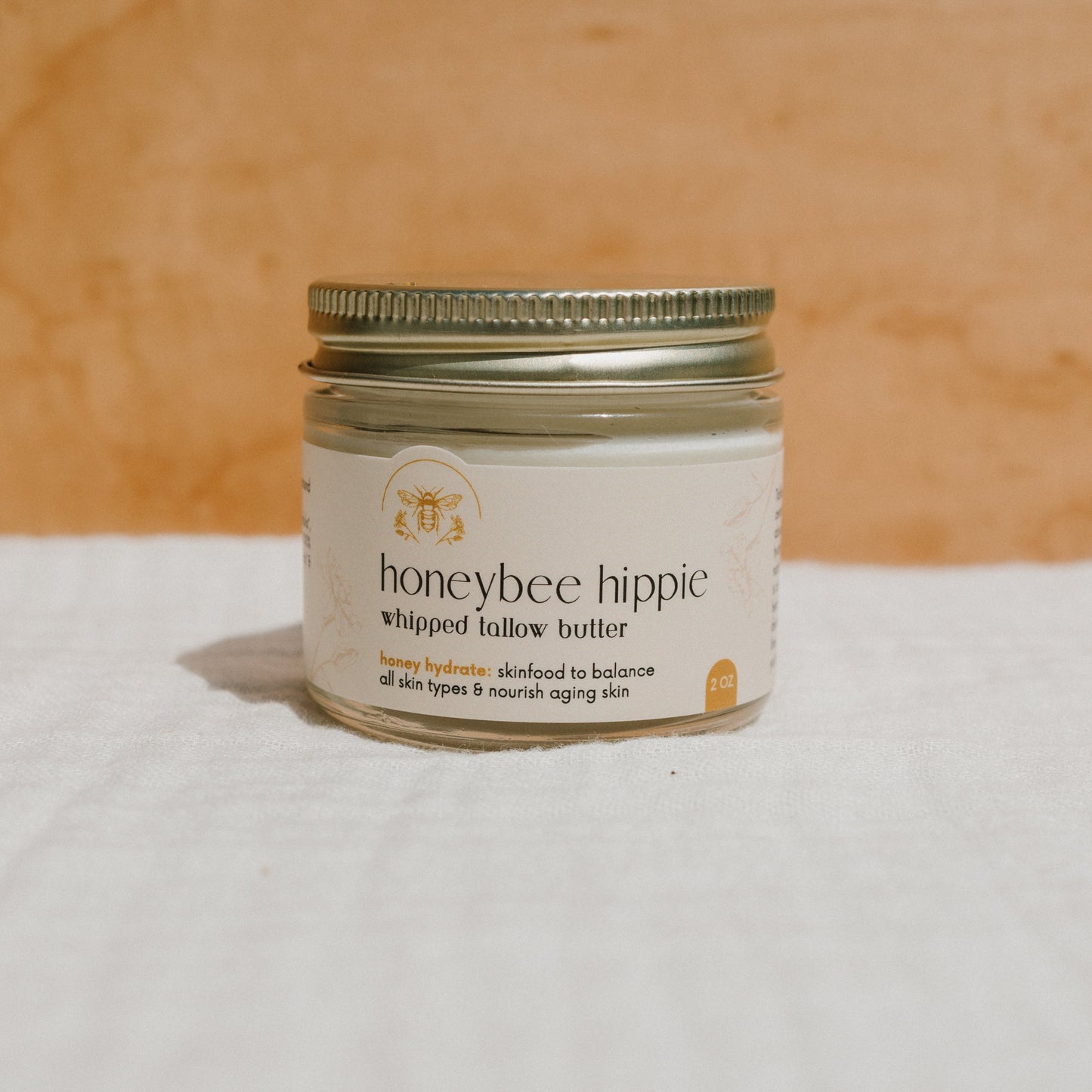 A glass jar with a metal lid contains "honey hydrate whipped tallow butter" by Honeybee Hippie, infused with Manuka honey. The label touts "hydrating and nourishing" benefits for all skin types, and the jar rests on a white cloth against a warm-toned background.