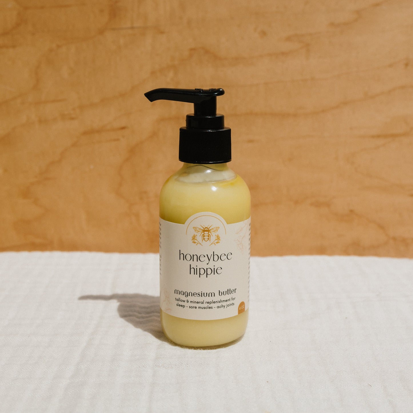 A yellow Honeybee Hippie magnesium butter with a black pump lid sits on light cloth, set against a wood-textured background highlighting its bee logo. Ideal for soothing sore muscles, aches, and stress relief, it offers comfort for both body and mind.