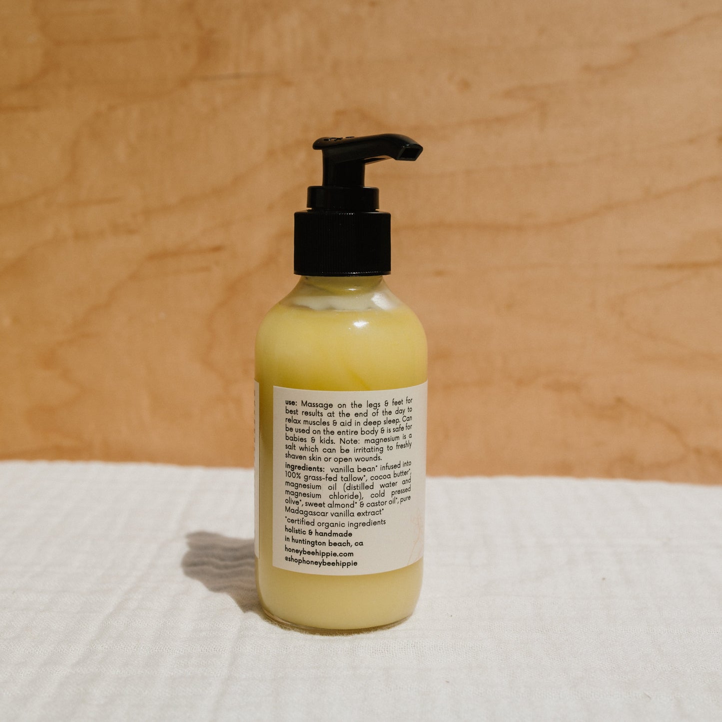 A yellow Honeybee Hippie Magnesium Butter bottle with a black pump rests on a white textured cloth, set against a wooden backdrop. The label highlights skin hydration and stress relief benefits, lists ingredients and usage instructions, and includes a website link at the bottom.