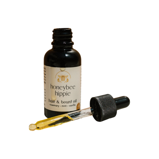 A black Honeybee Hippie hair & beard oil dropper bottle is shown, featuring organic oils. The golden oil dropper is placed in front, highlighting rosemary, mint, and vanilla on the label—ideal for healthy hair growth.