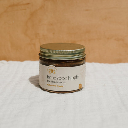 A jar of Honeybee Hippie's herbal infused raw honey mask with a gold lid sits elegantly on a white surface. The label, set against a wooden background, promises hydration and detoxification for rejuvenating skin.