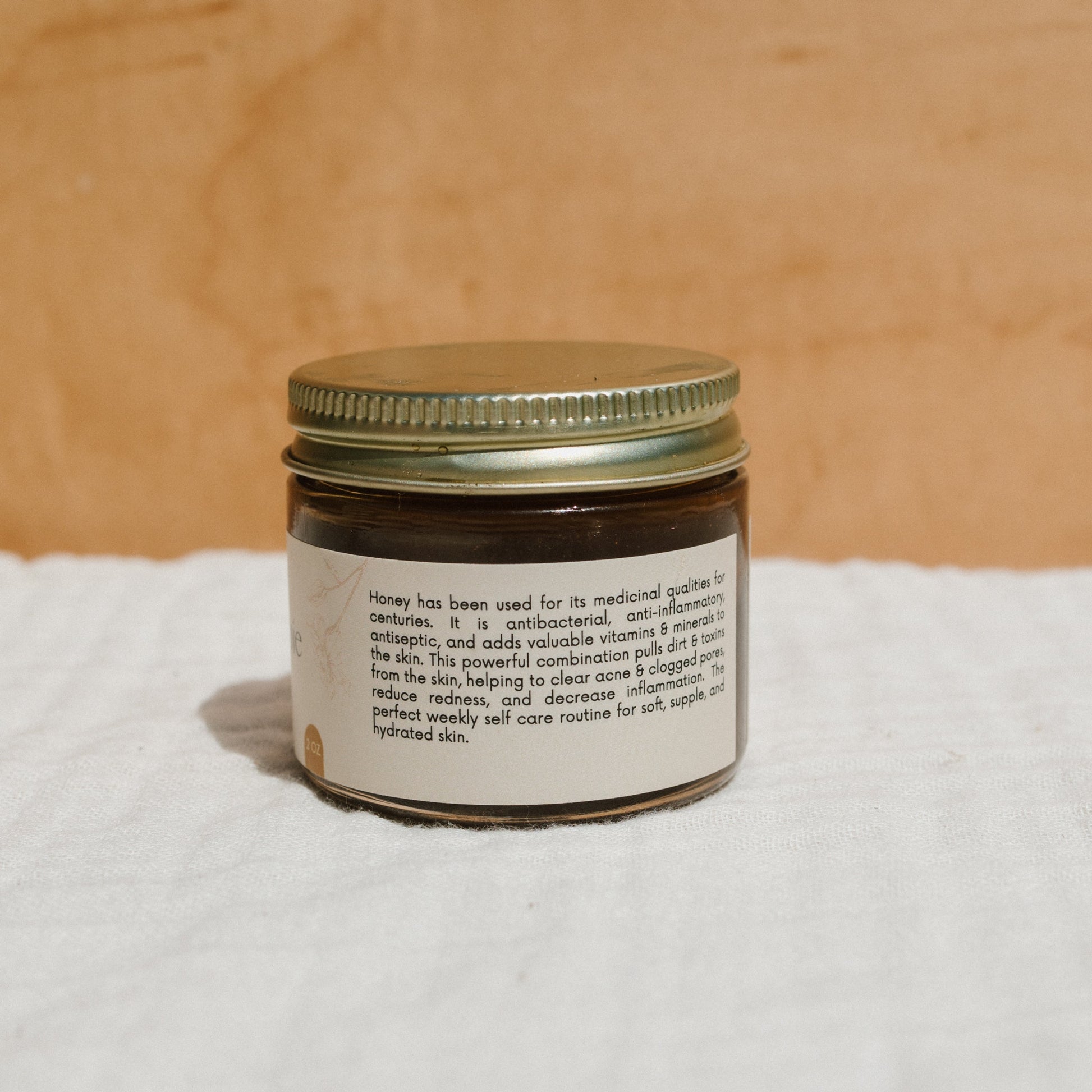 A jar with a metal lid holds Honeybee Hippie's "herbal infused raw honey mask" on a white surface, showcasing its detoxifying and antiseptic benefits for skin hydration. The warm beige background enhances its natural charm.