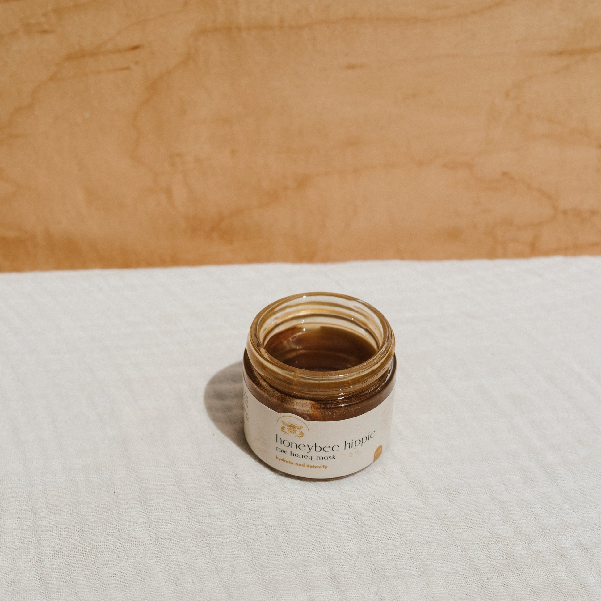 A small glass jar labeled "Honeybee Hippie" sits open on a textured white surface. The jar contains a honey-colored substance: the herbal infused raw honey mask for skincare. The light wooden background enhances its organic appeal.