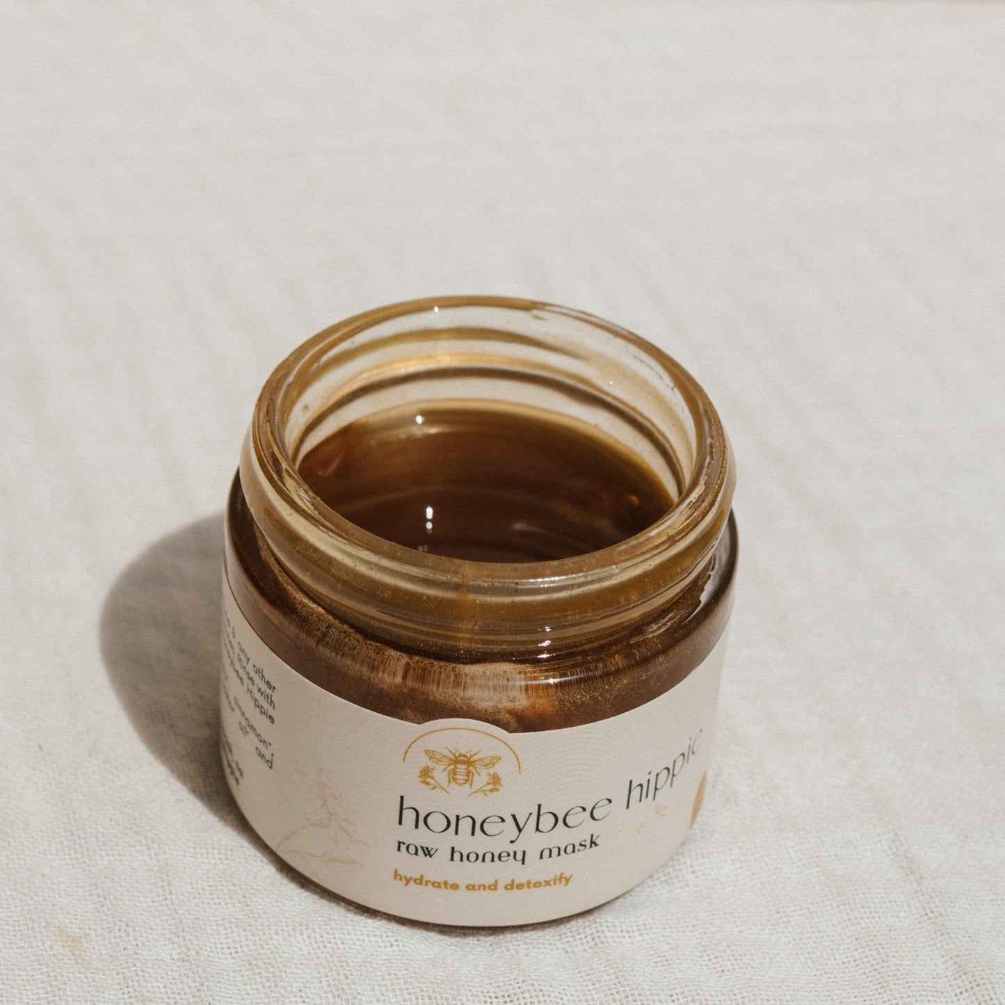 Open a jar of Honeybee Hippie's herbal infused raw honey mask on a textured surface. This detoxifying skincare essential is filled with golden-brown substance, labeled "hydrate and detoxify.