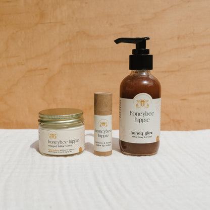 The Honeybee Hippie "honey bundle," featuring hibiscus and honey lip butter, a honey glow lip balm in a cardboard tube, and body wash in a pump bottle, is displayed against a wooden background.