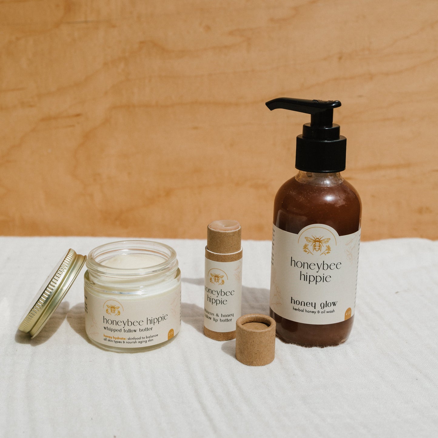 The Honeybee Hippie's Honey Bundle, featuring whipped tallow butter, two skin stick tubes, and a honey glow lotion pump bottle on a cloth surface against a wooden backdrop, offers moisturizing benefits for optimal skin health.