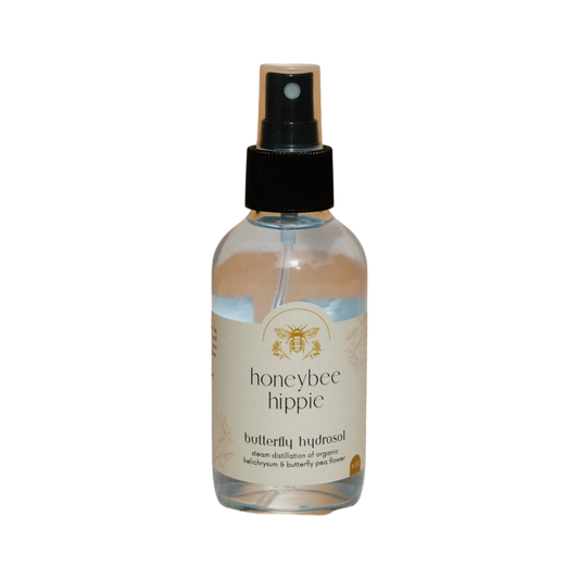 A spray bottle labeled "butterfly hydrosol" by Honeybee Hippie stands against a black background. The label highlights its organic blend of Butterfly Hydrosol and Helichrysm, capturing the delicate aroma of butterfly pea flower with a bee logo.