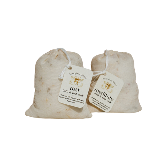 Two beige cloth bags with string ties feature tags that read "Rest bath & foot soak" and "Meditate bath & foot soak." Adorned with a bee logo and organic herb details, these Honeybee Hippie products promise relaxing sleep against a crisp white background.