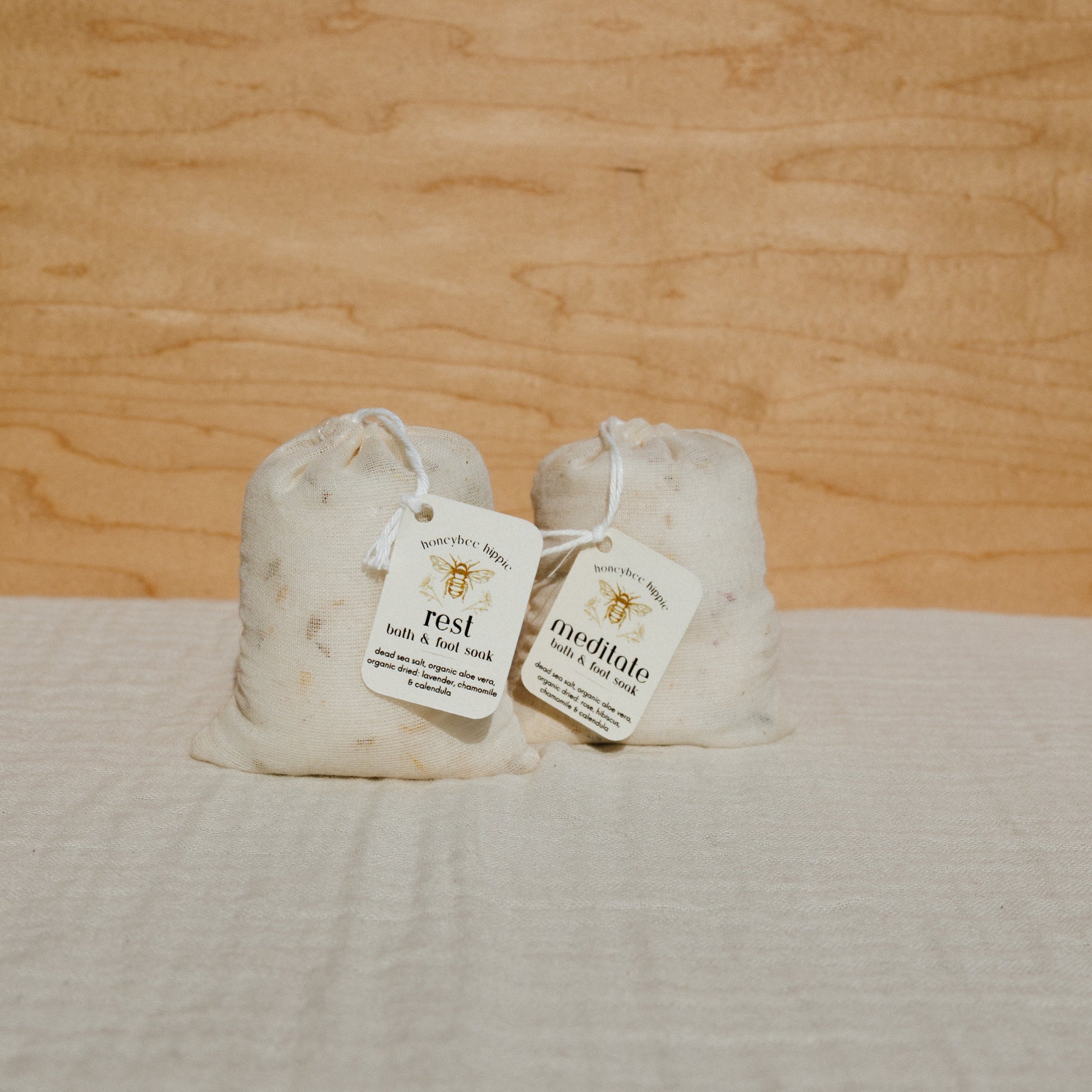 Against a wooden background, two labeled sachets of organic herb bath soak sit on light woven fabric. One reads "rest" from Honeybee Hippie and the other reads "meditate" by Birchtree Apothecary, both ideal for a relaxing sleep.