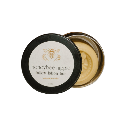 A round tin with an open lid reveals a Honeybee Hippie tallow lotion bar made from grass-fed tallow, featuring a bee logo and "tallow lotion bar, hydrate & soothe, 2 oz." on the lid. The moisturizer is adorned with a decorative bee imprint.