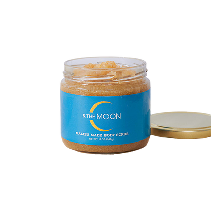 A transparent jar contains the Malibu Made Body Scrub, a light brown blend of natural vanilla brown sugar and organic oils. The blue label reads "C & The Moon." The lid is off and placed next to the jar, revealing its 12 oz (340g) contents.