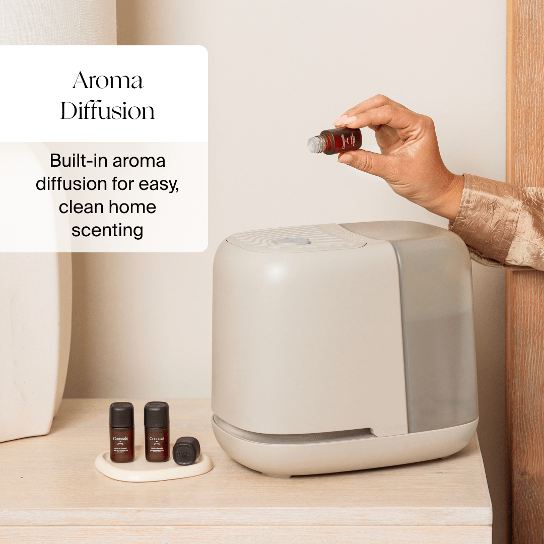 A hand adds oil to a white Bedside Humidifier 2.0 by Canopy on a beige surface. Three oil bottles sit on a small tray beside the diffuser. Text reads: "Aroma Diffusion: Built-in aroma diffusion for effortless, clean home scenting with simplicity and elegance.