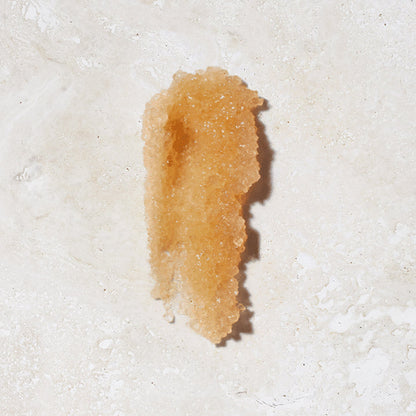 A light brown, granular substance that appears to be Malibu Made Body Scrub by C & The Moon is spread on a white surface. The scrub forms an irregular shape with a shadow falling to one side.