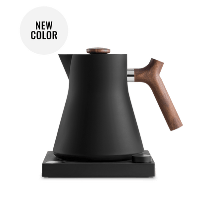 The Corvo EKG Electric Kettle by Fellow Wholesale, with its sleek matte black finish and wooden handle and knob, is elegantly perched on its square black base. Precision engineered, it boasts variable temperature control. The image emphasizes the kettle's minimalist design and includes a white circle noting "NEW COLOR.