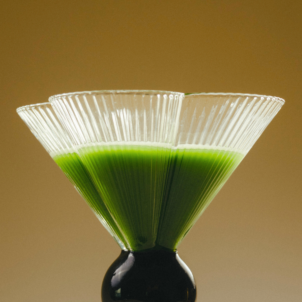 Three conical glasses with ridged patterns, filled halfway with vibrant green ceremonial-grade matcha, are arranged overlapping each other against a muted brown background. The base is a black spherical stand supporting the arrangement, highlighting the energy and metabolism-boosting properties of LAKA's LAKA Matcha™ Sticks.