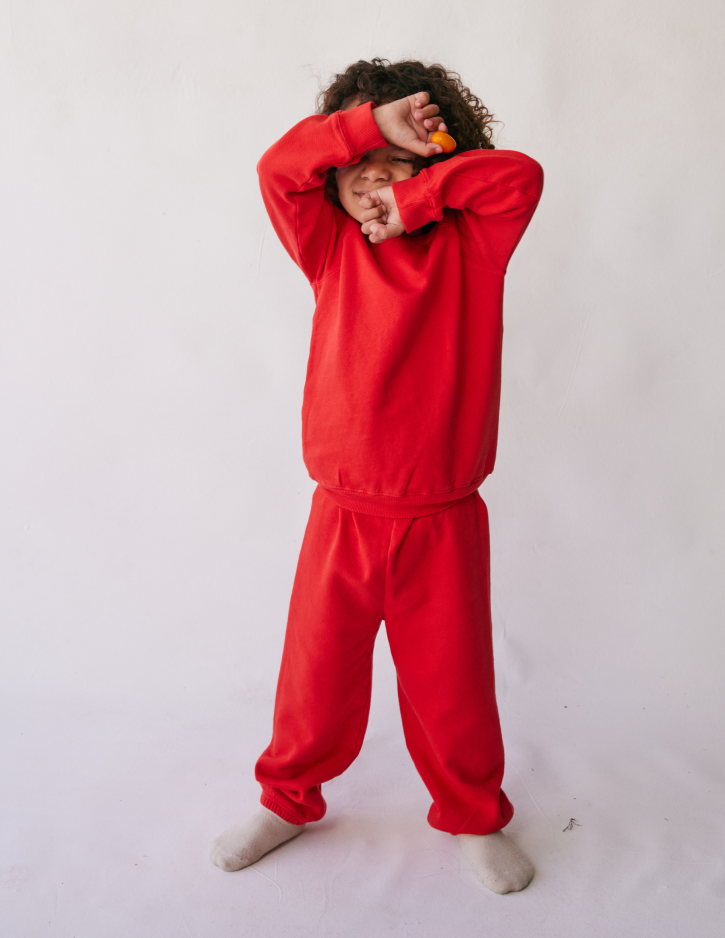 A young child dressed in a cozy outfit featuring "The Lil' Classic Sweatpant" by all the babies and a red sweatshirt stands against a plain white background. They have curly hair and are playfully covering their face with their arms while standing barefoot.