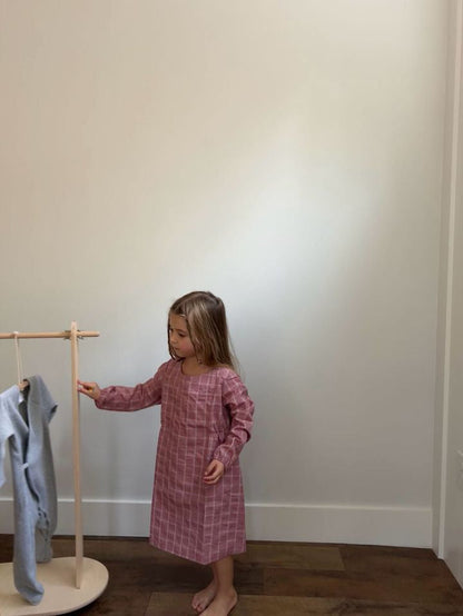 A young girl in "the DAY DRESS plaid" by cabane childrenswear, featuring long sleeves and a charming raspberry plaid design handmade in Vancouver, stands barefoot on a wooden floor. She holds a stick near a small wooden rack with clothes hanging on it. The room's light-colored walls enhance the appeal of this limited edition attire.