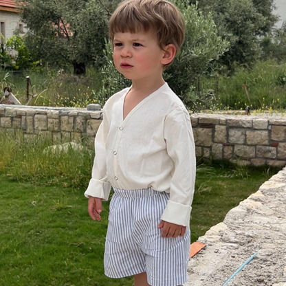 A young child stands outdoors on a grassy area beside a stone wall, wearing the TAKE CARE TOP raw silk blouse by cabane childrenswear with rolled-up sleeves and striped shorts, looking slightly to the side. The background features trees and a house with part of a red roof visible.