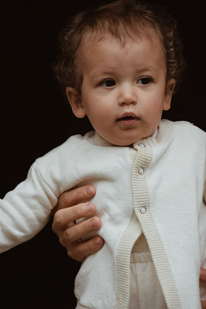 A toddler with curly hair is wearing the Soul Sweater Baby + Kid, a white, ethically handmade knit cardigan by cabane childrenswear. An adult's hand is gently supporting the child from the side against a dark background. The child looks slightly to the right with a calm expression.
