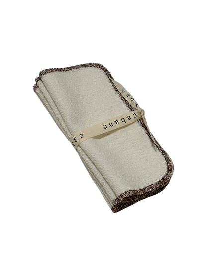 A beige french terry washcloth with brown edges, tied with a cream ribbon labeled "cabane," handmade in Vancouver from an organic cotton/hemp blend. Displayed stylishly on a black background, this ORGANIC WASHCLOTH by cabane childrenswear exemplifies elegance and eco-friendliness.