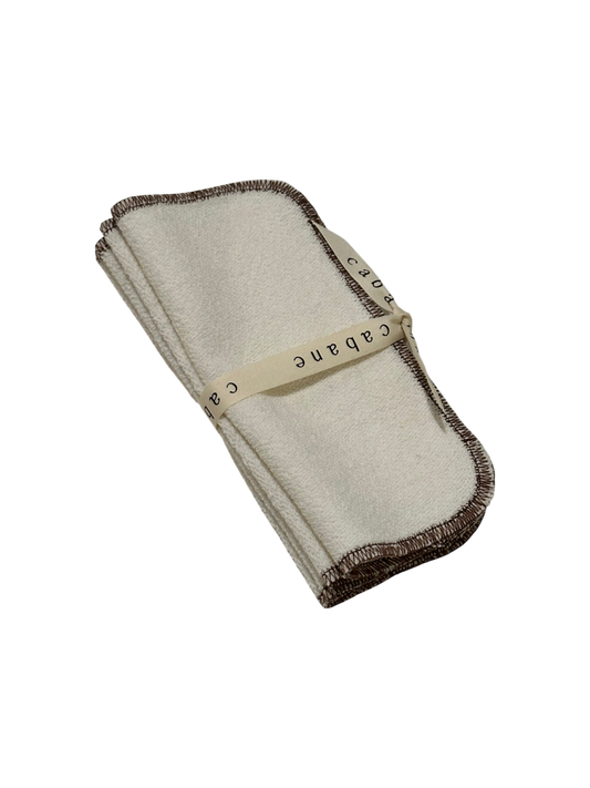 A beige french terry washcloth with brown edges, tied with a cream ribbon labeled "cabane," handmade in Vancouver from an organic cotton/hemp blend. Displayed stylishly on a black background, this ORGANIC WASHCLOTH by cabane childrenswear exemplifies elegance and eco-friendliness.