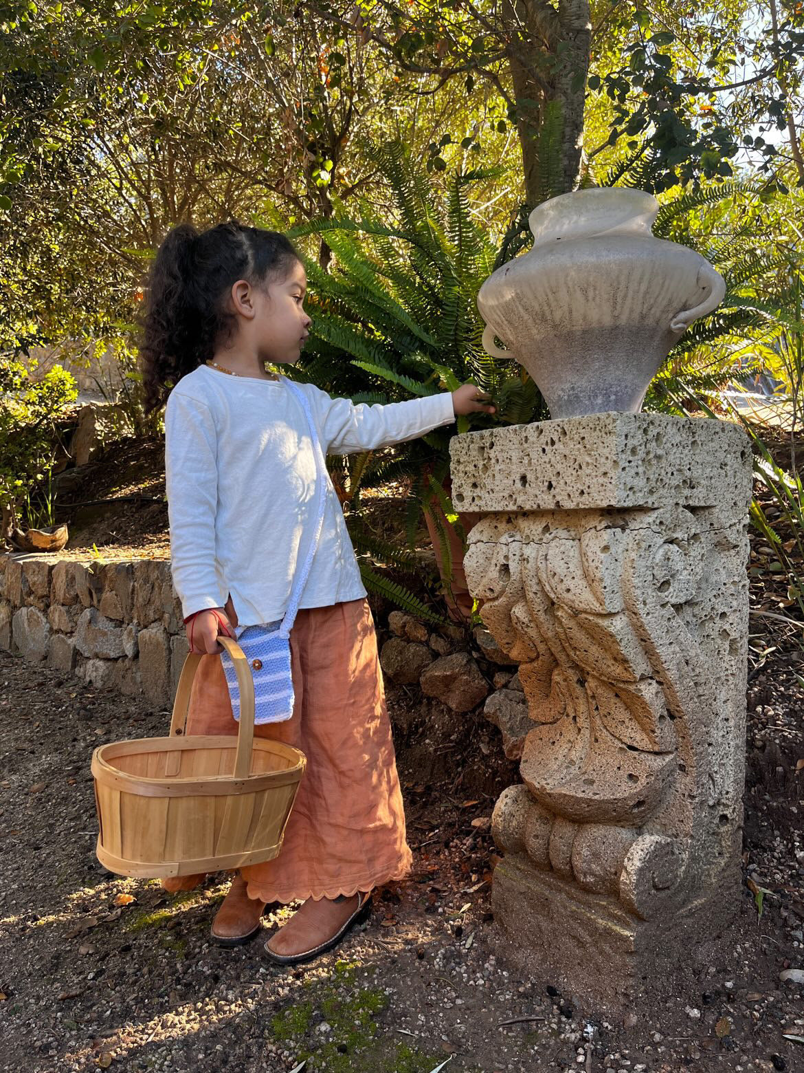 A young girl with curly hair, wearing a RESORT TOP hemp jersey from cabane childrenswear and rust-colored pants, stands outdoors holding an empty wooden basket. She reaches out to touch a stone structure adorned with a decorative urn amidst a greenery-filled garden, showcasing the charm of handmade in Calgary fashion.